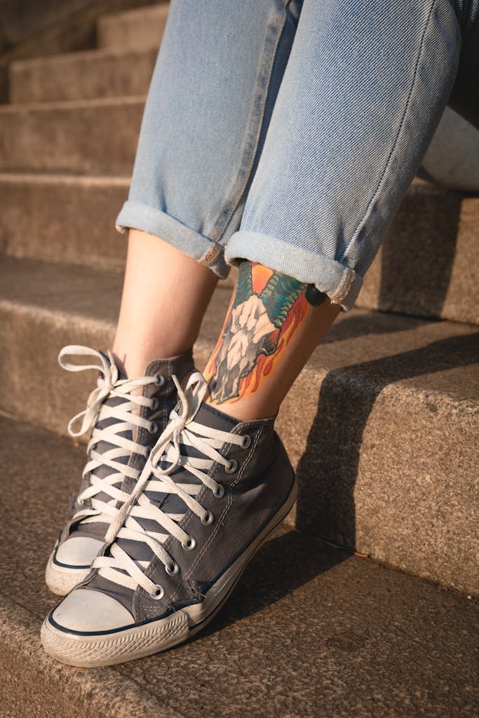 Close-up of a woman's legs in jeans and sneakers, showcasing a colorful leg tattoo in an urban setting.