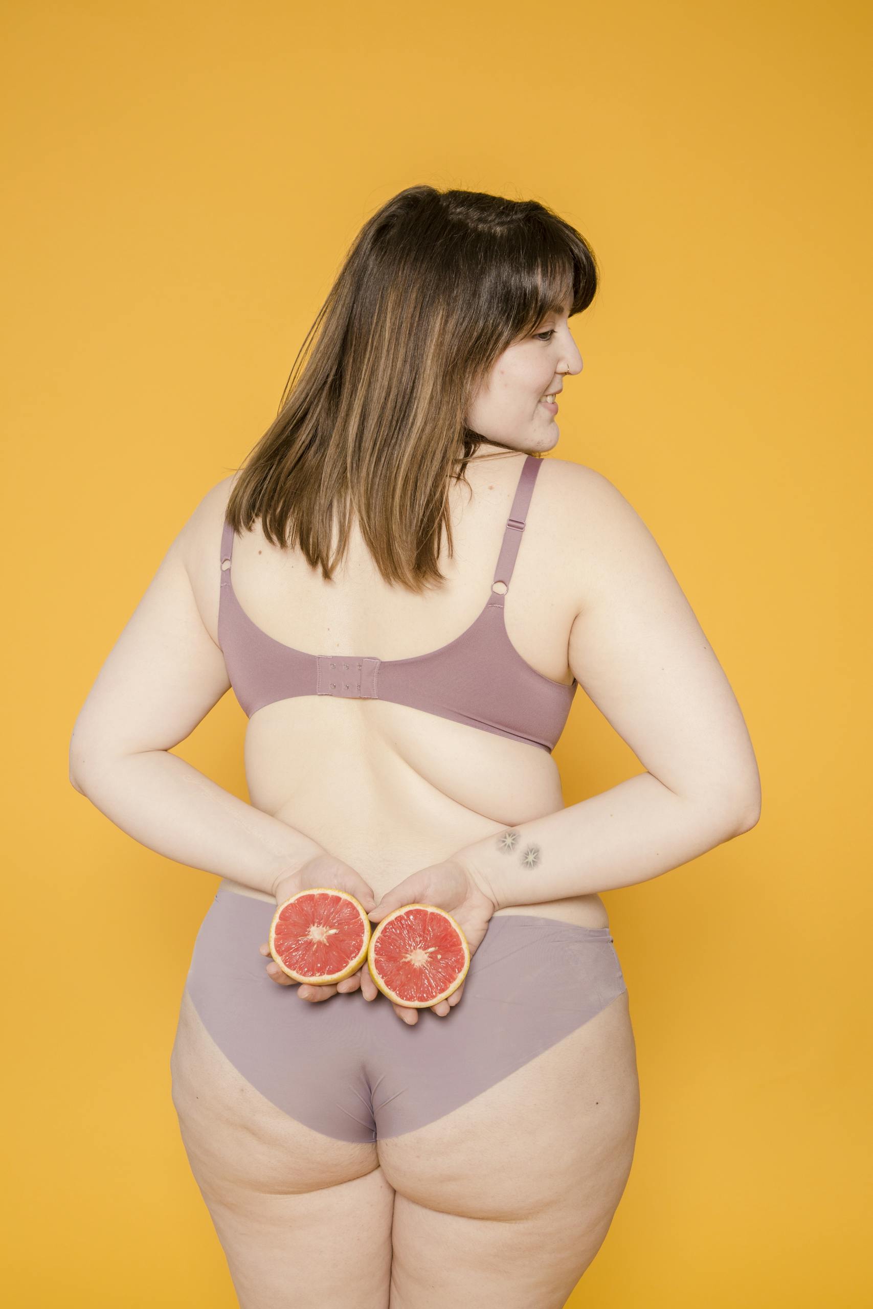 Plus size woman holding grapefruits behind her back, smiling against a bright yellow background.